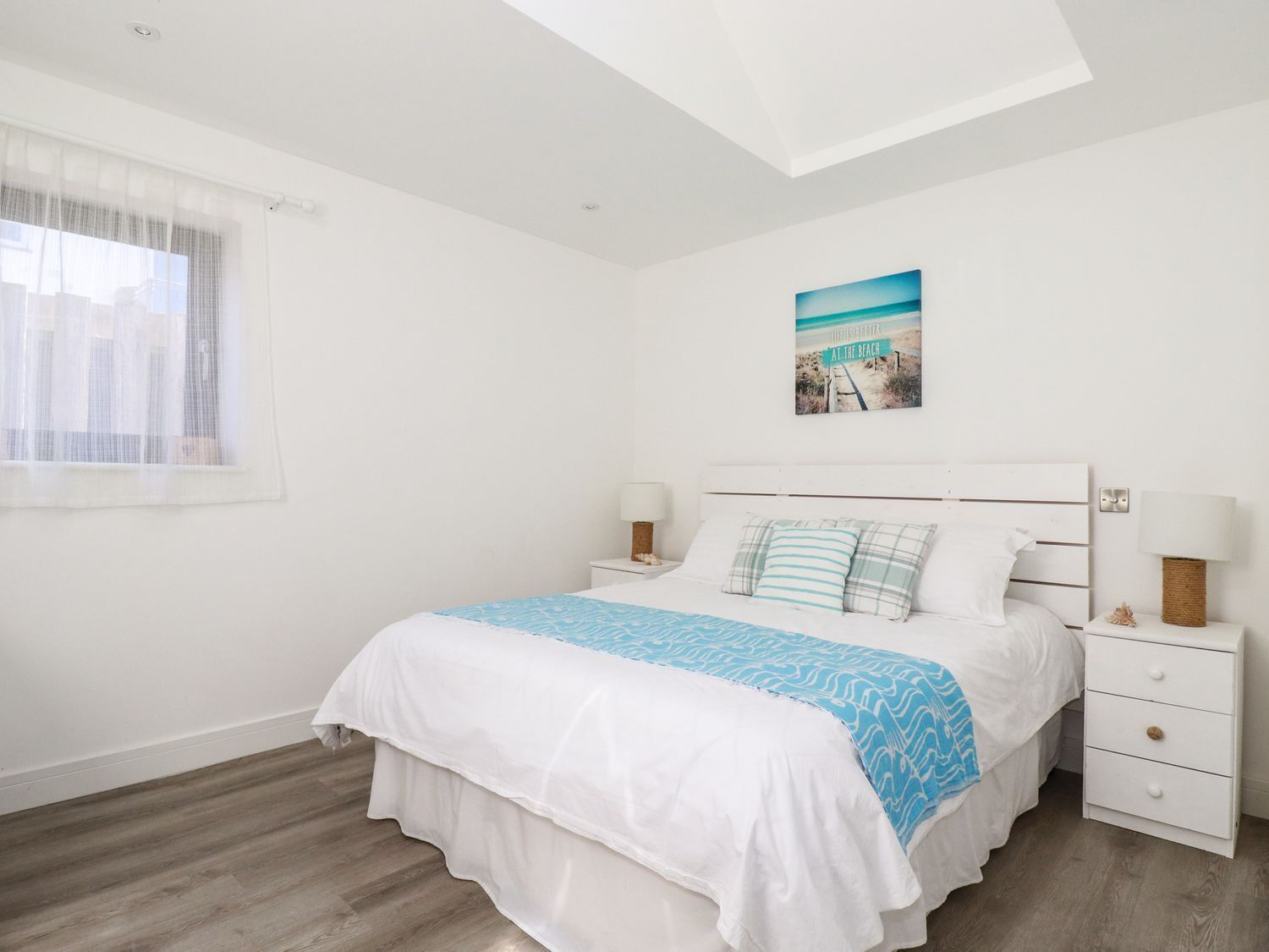 Un dormitorio con una cama, dos mesitas de noche, una ventana con cortinas translúcidas, una lámpara de techo y una decoración mural con temática playera.