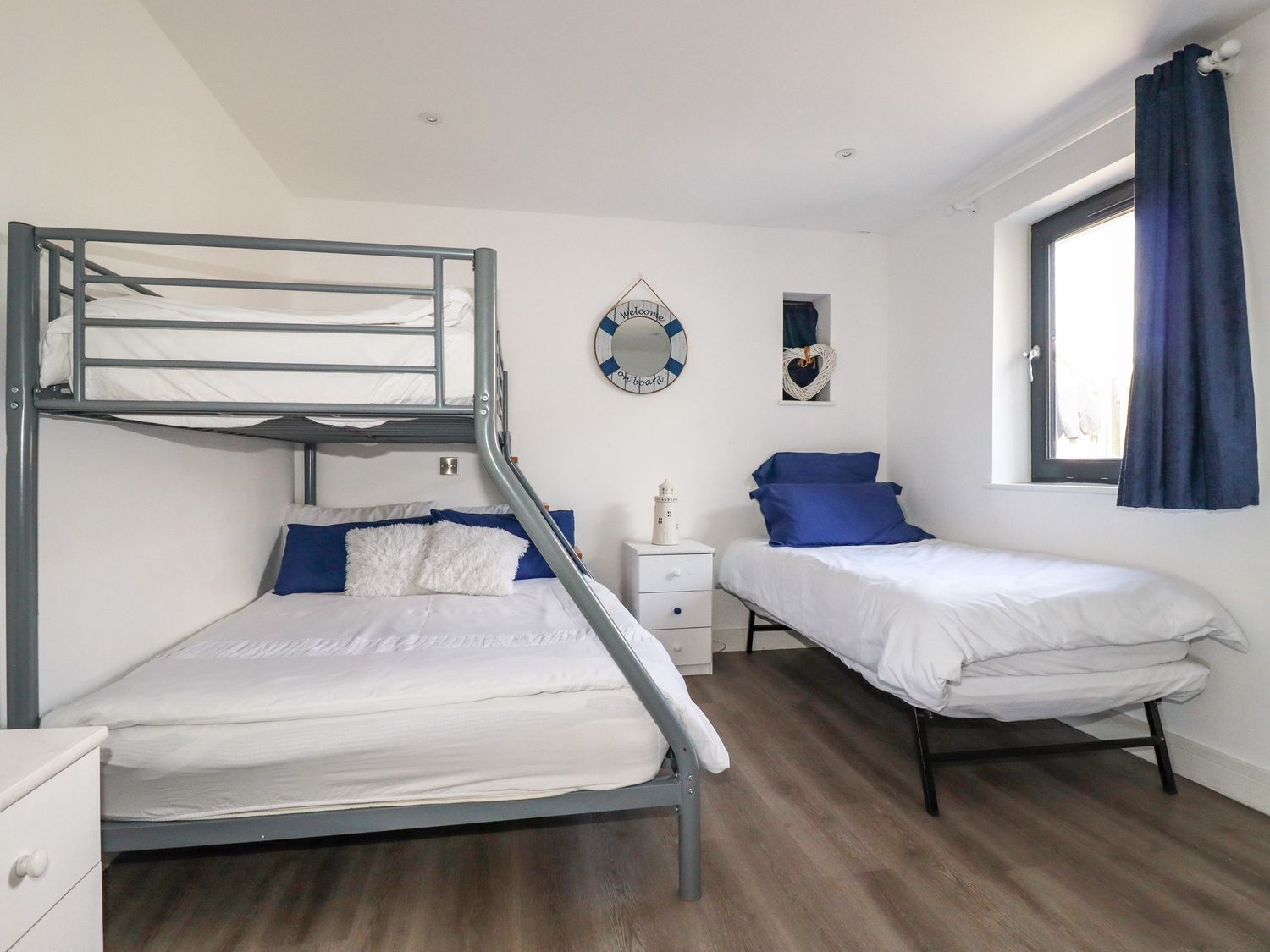 A dormitory room with bunk beds, a small bedside table, a wall clock, and a window with curtains.