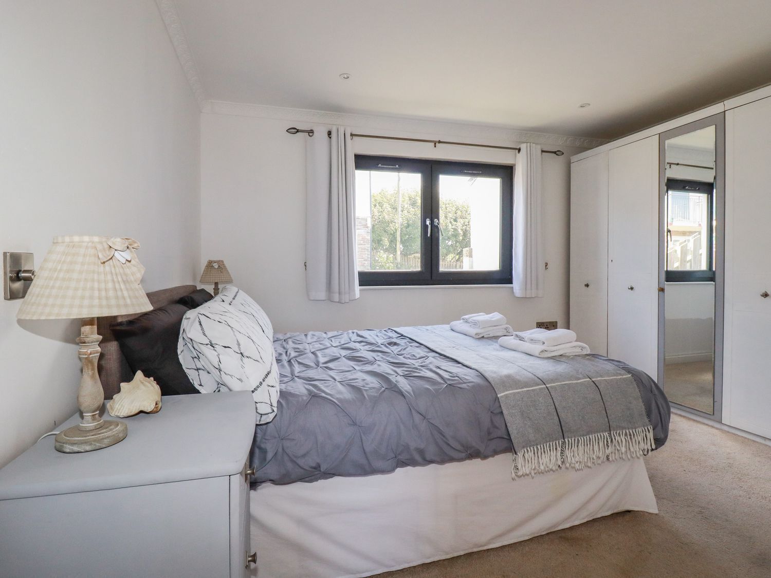 A bedroom with a bed, a bedside table with a lamp, a mirror, and a window with curtains.