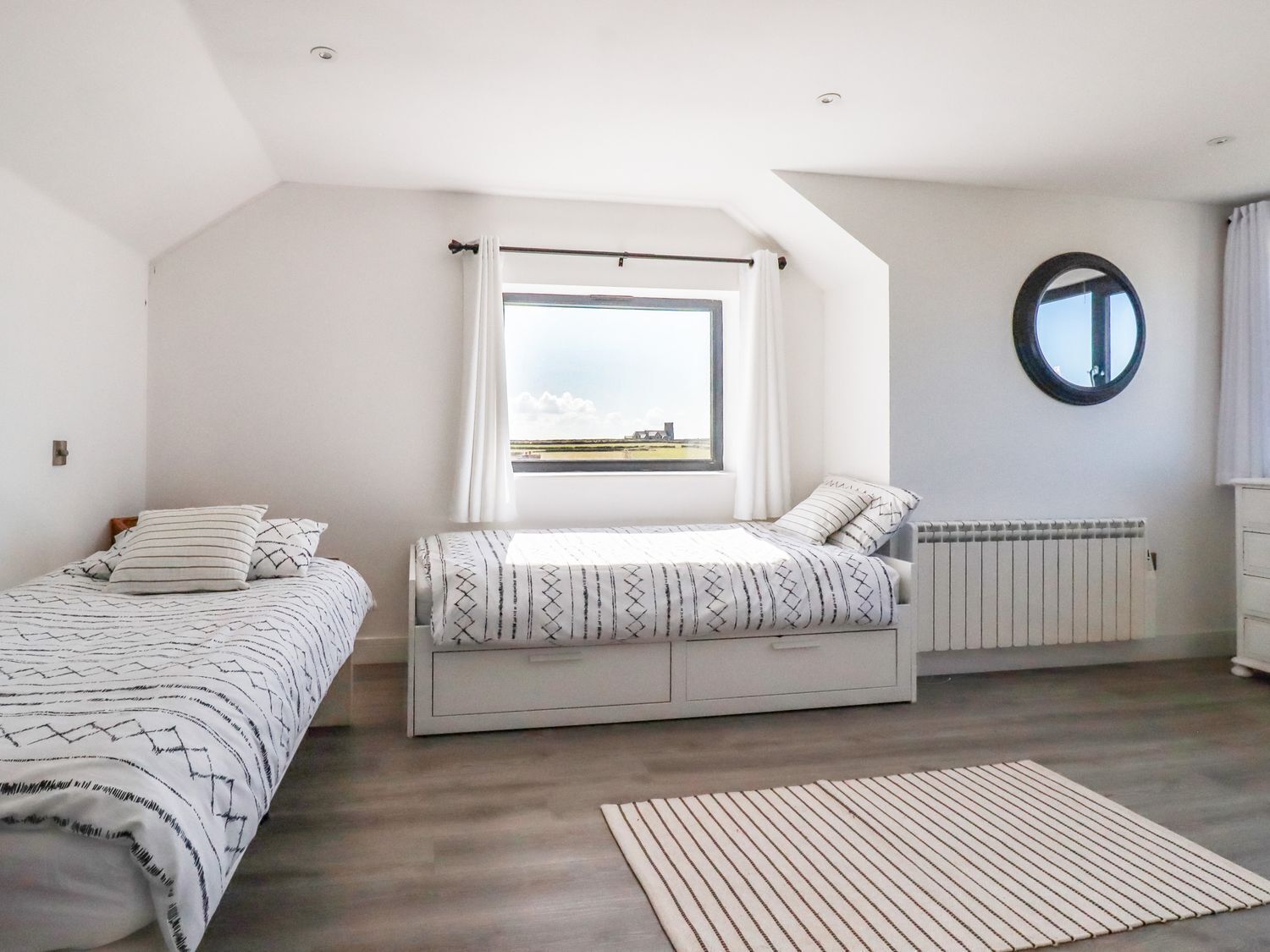 Un dormitorio con dos camas, una ventana con vistas, un radiador y un espejo circular en la pared.