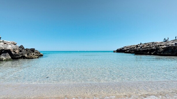 Una playa con agua cristalina, afloramientos rocosos y algunas personas a lo lejos.