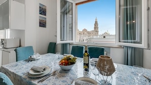 Comedor con mesa lista para dos, un cuenco de fruta, una botella de vino y vista a un edificio histórico a través de amplios ventanales.