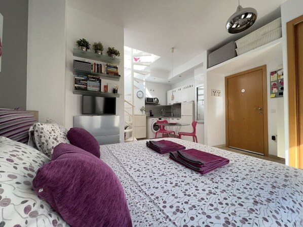 Un dormitorio con cama, mesita de noche, estantería, escritorio y kitchenette.