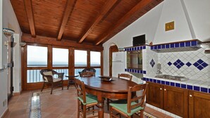 A dining area with a wooden table and chairs, a kitchen with blue and white tiles, and a view of the sea through large windows.