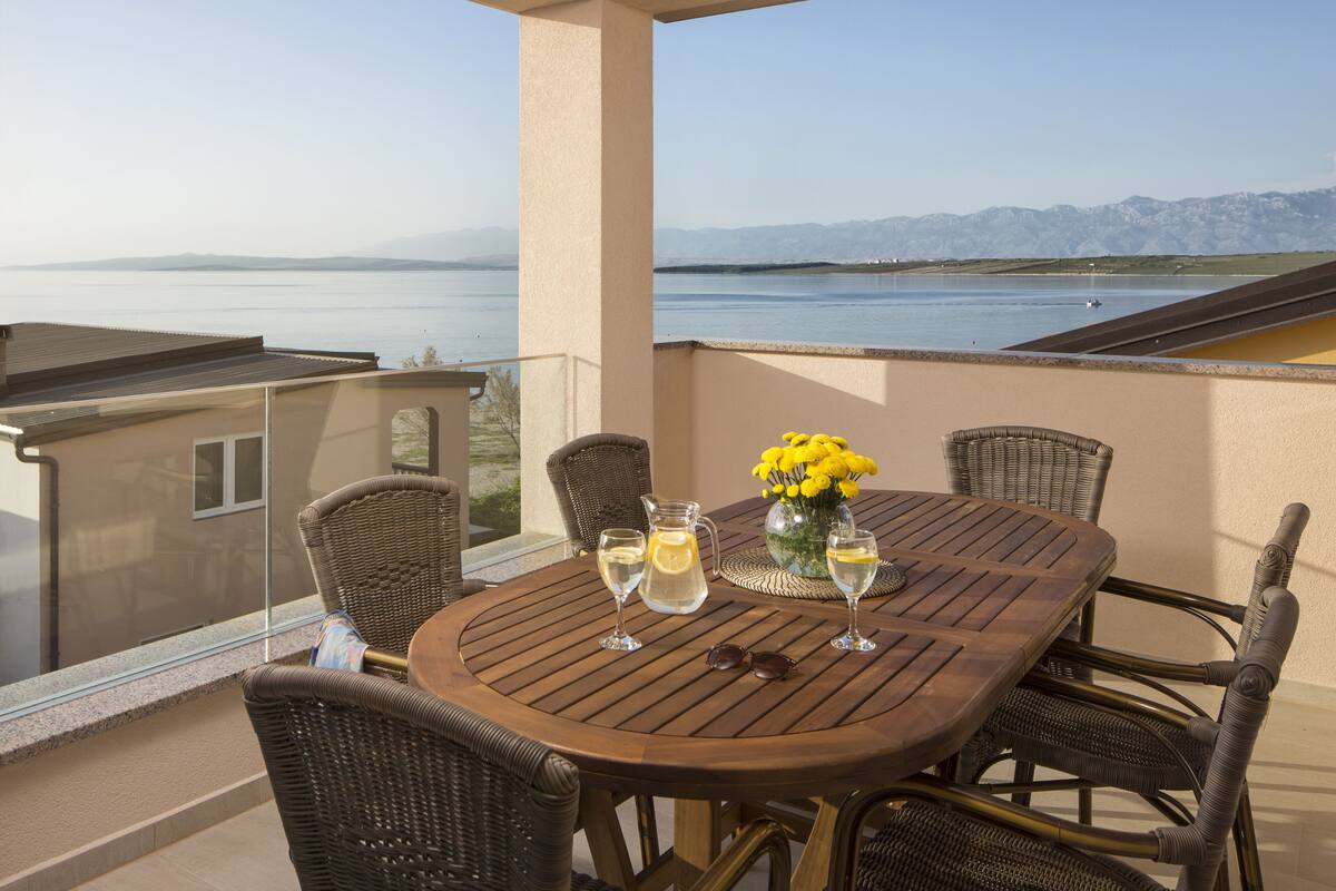 A round table with wicker chairs on a balcony overlooking a body of water and mountains.