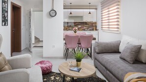 A modern living room with a sofa, a coffee table with a book and a plant, a dining area with pink chairs, and a kitchen in the background. - Apartments Vista Exclusive  (Vir)