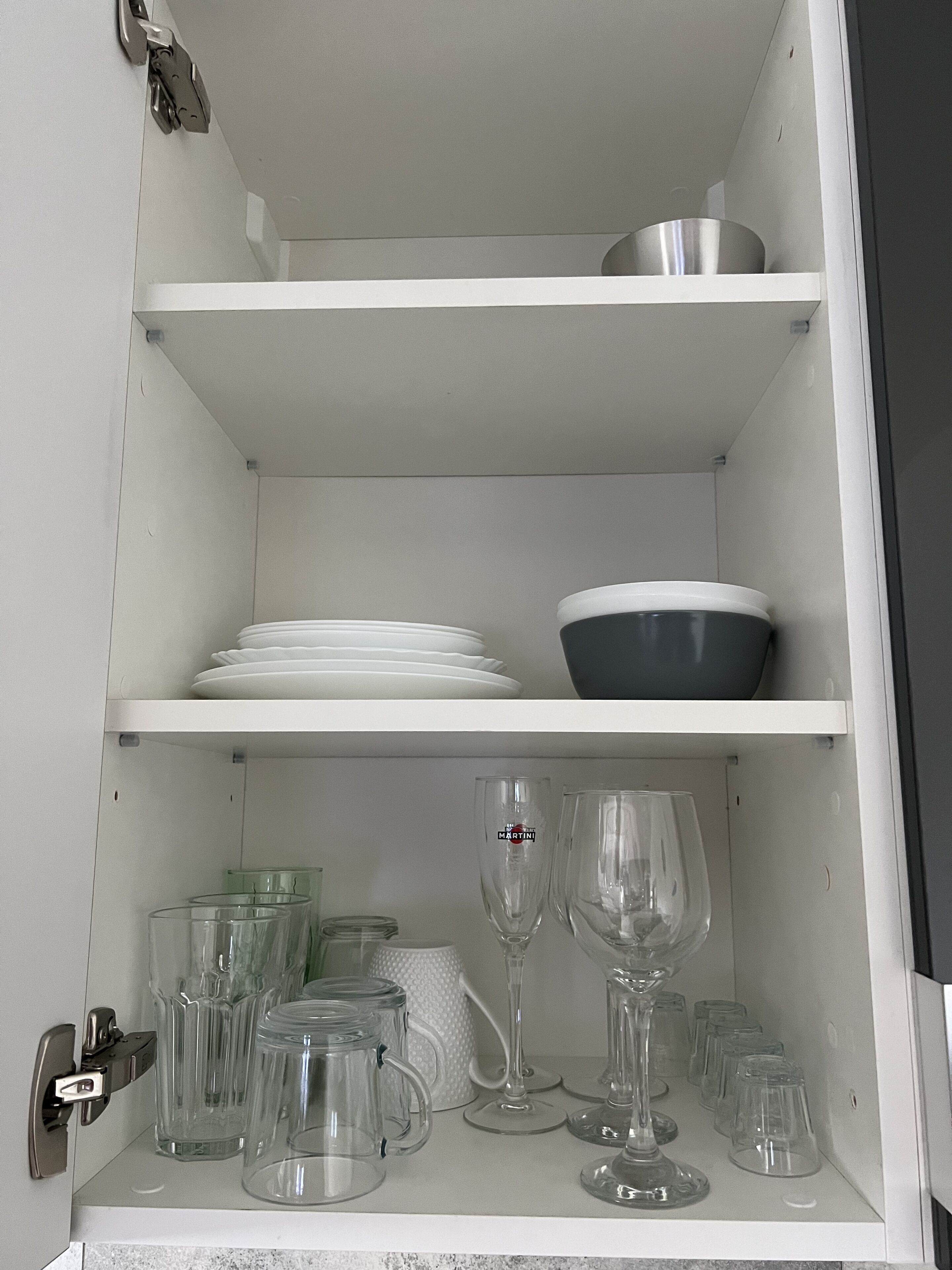A kitchen cabinet with white shelves, holding plates, bowls, and various glassware.