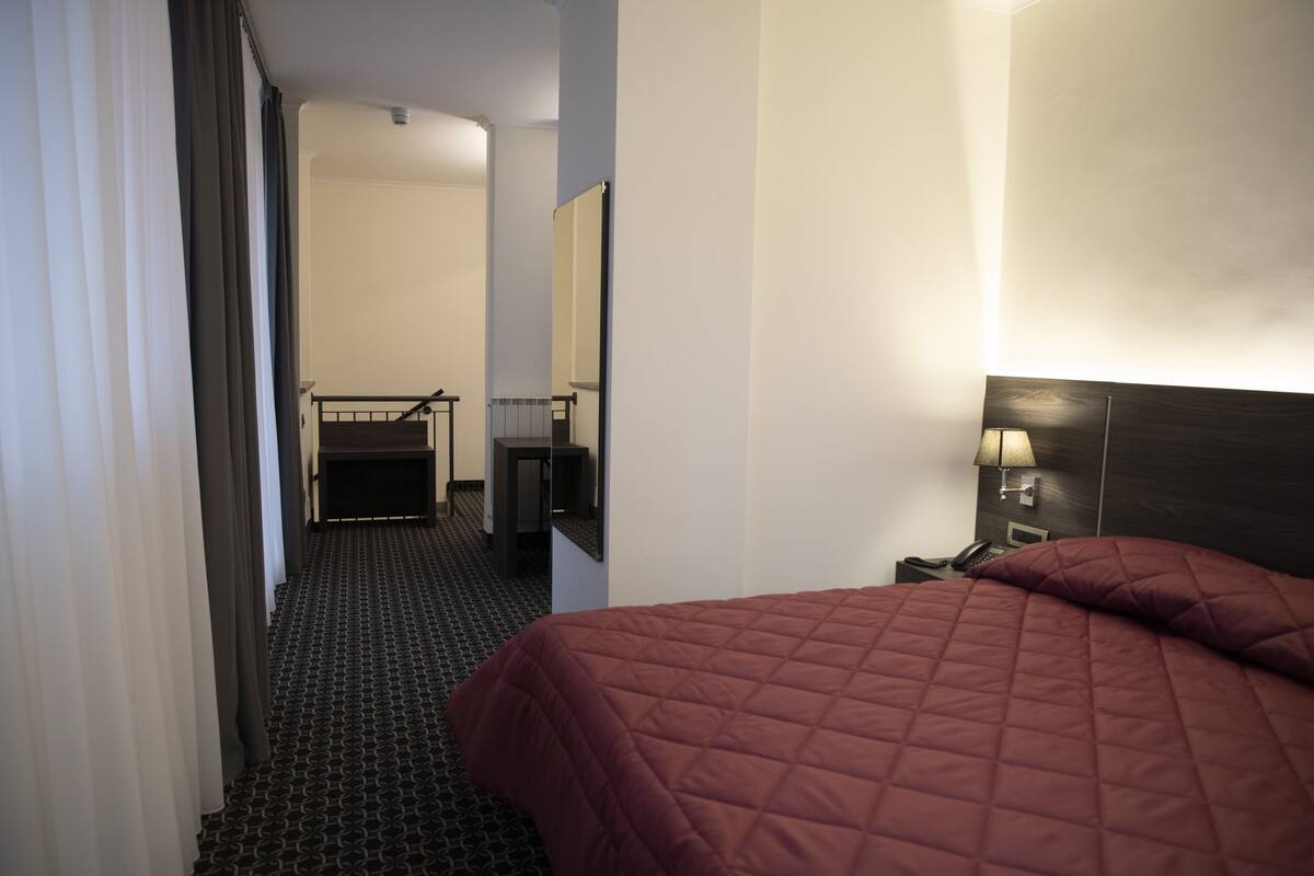 A hotel room with a bed, a desk, a chair, a mirror, and a nightstand with a lamp.
