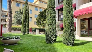 A courtyard with neatly trimmed grass, tall coniferous trees, and a building with a red awning. - Gemelli Hotel (Rome)