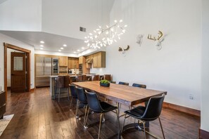 A spacious dining area with a long wooden table and modern chairs, a kitchen in the background, and decorative antlers on the wall.