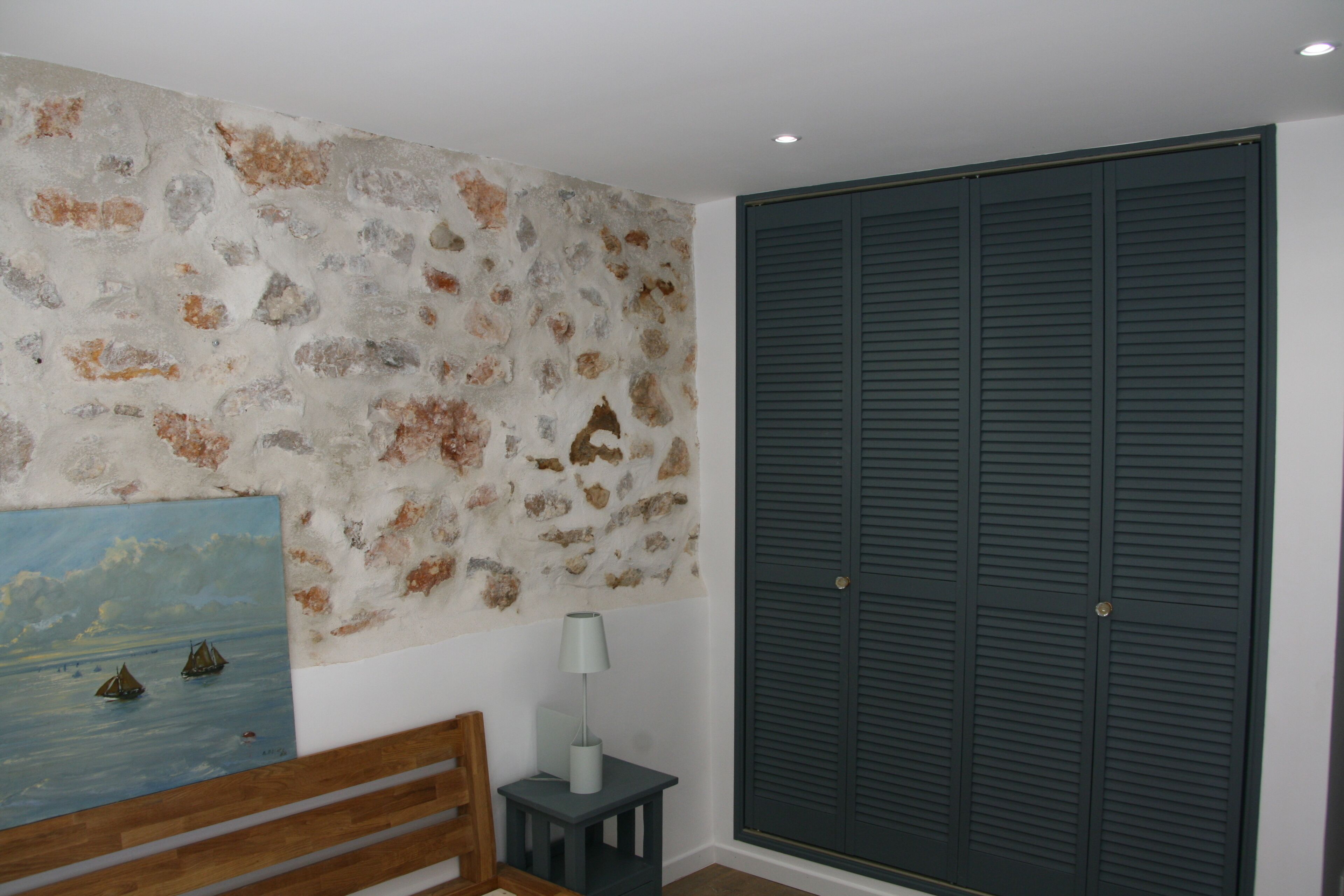 A room with a stone wall, a painting, a wooden bench, a small table with a lamp, and a large dark closet.