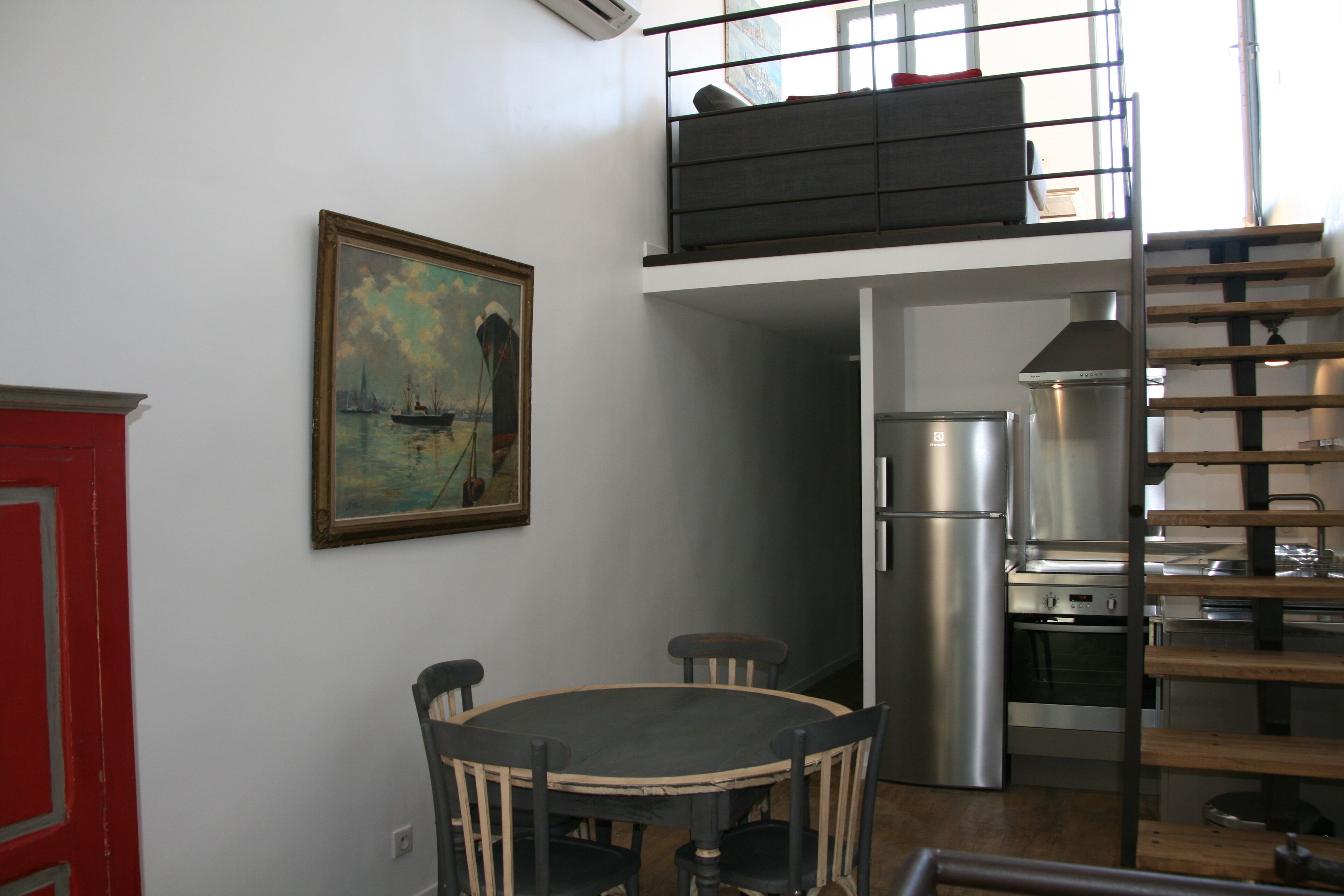 A dining area with a round table and chairs, a painting on the wall, a kitchen with modern appliances, and a staircase leading to an upper level.