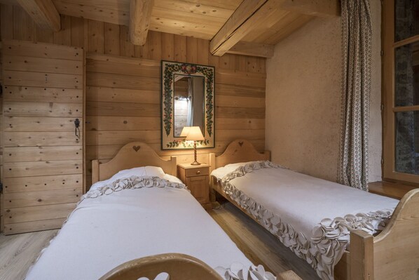 Habitación de cabaña de madera con dos camas individuales, una mesita de noche con lámpara, un espejo con marco floral y una ventana con cortinas.