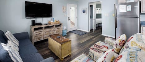 Un salón con televisor de pantalla plana, mueble bajo de madera, sofá azul y otomana estampada.