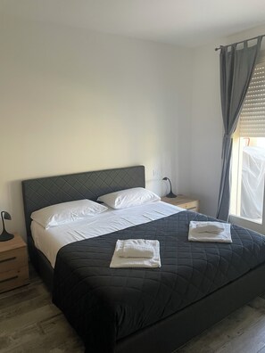 Een keurig opgemaakt bed met een donkere quilt en witte lakens, een bedlamp en een houten nachtkastje.
