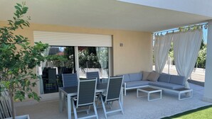 Un patio exterior con un sofá blanco, dos sillones y una mesita. Hay una planta en maceta y una puerta de cristal que da al interior.