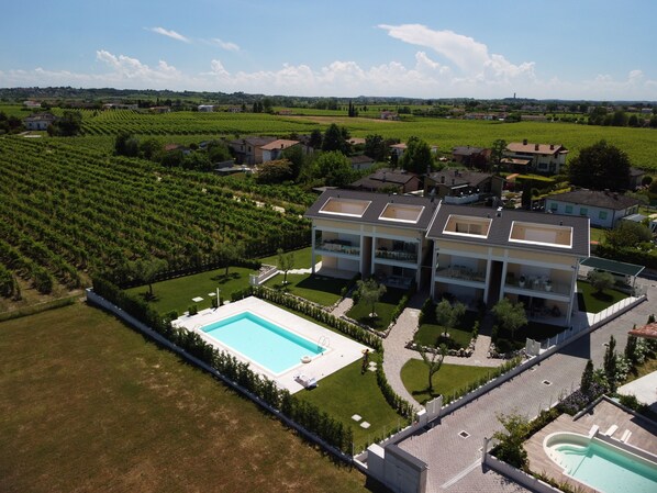 A modern multi-story building with a swimming pool, surrounded by lush green fields and vineyards.