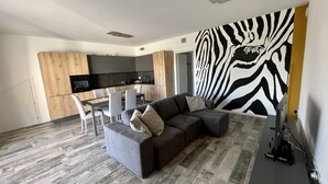 Una sala moderna con un sofá seccional gris, una mesa de comedor con sillas, una zona de cocina con armarios oscuros y un mural de cebra en la pared.
