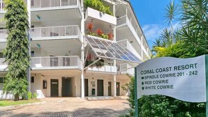 Un edificio de varios pisos con balcones, un letrero que dice 'CORAL COAST RESORT' y un área de entrada pavimentada.