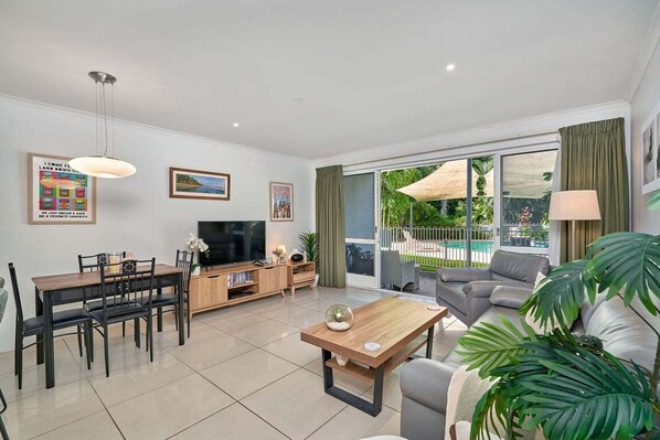 A modern living room with a dining area, a flat-screen TV, and a large window with a view of greenery.