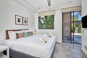 Relax in a spacious room with a large bed, bedside table, and flat-screen TV, offering a view of lush greenery through a large window.