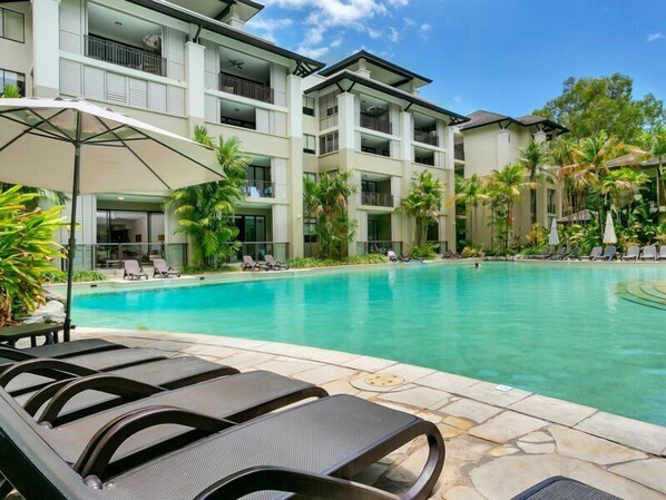 Relax by the pool surrounded by lounge chairs and umbrellas, with a multi-story building in the background.