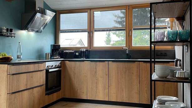 A modern kitchen with wooden cabinets, a built-in oven, a stainless steel range hood, and a window overlooking a residential area. - Lovingly restored vacation apartment in alpine chic style DG (Ottobeuren)