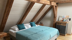 A bedroom with a sloped ceiling, wooden beams, a bed with teal bedding, a nightstand, a cabinet with artwork, and a cowhide rug. - Lovingly restored vacation apartment in alpine chic style DG (Ottobeuren)