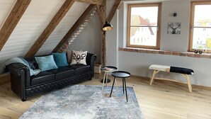 A loft-style room with exposed wooden beams, a black leather sofa, a small round table, and a bench. - Lovingly restored vacation apartment in alpine chic style DG (Ottobeuren)