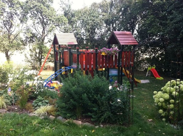 A playground with a wooden play structure, slides, and climbing areas surrounded by greenery.