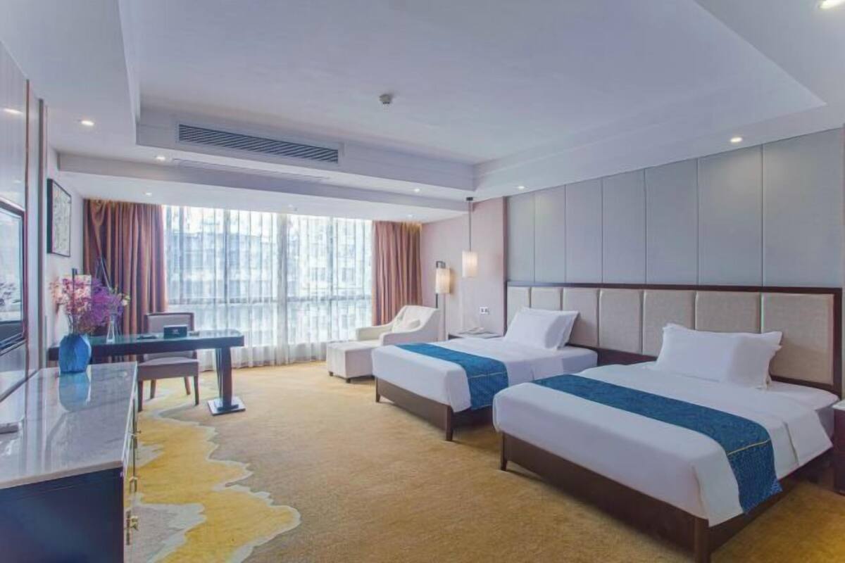 A hotel room with two beds, a desk, a chair, and a large window with curtains.