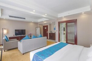 Habitación de hotel con cama, sofá, escritorio con televisor y baño visible a través de una puerta de vidrio.