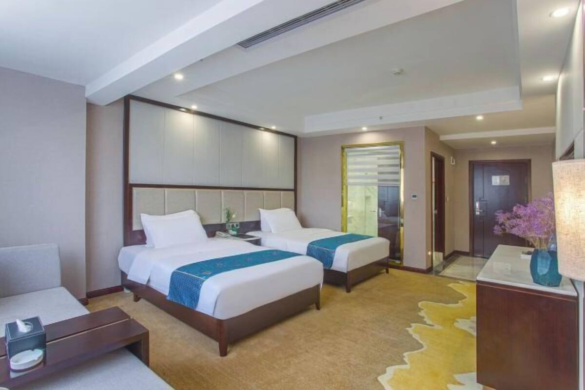 A hotel room with two beds, a desk, a chair, a nightstand, a lamp, and a window with blinds.