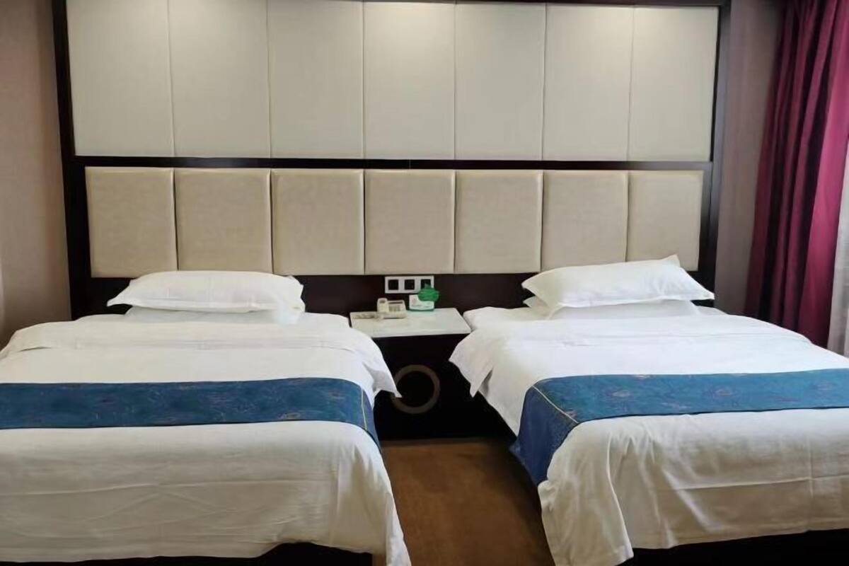 A hotel room with two beds, a large padded headboard, a small table with a phone and a green object, and a red curtain.