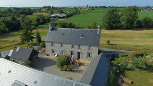 A stone building with a sloped roof, a paved courtyard, and a view of a green field and distant buildings. - Old farmhouse in Montchaton Orval sur sienne near Mont Saint Michel (Montchaton Orval sur sienne)