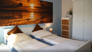 A bedroom with a bed, two pillows, a nightstand, a lamp, a large painting of a sunset over water, a plant on a shelf, and a white wardrobe. - Luxurious FH near the dike, sauna, fireplace, garden, beach chair (8 adults & baby) (Butjadingen)