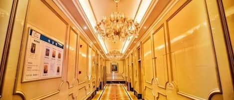 Un couloir d’hôtel avec un sol à carreaux, des lustres et un panneau d’information fixé au mur.