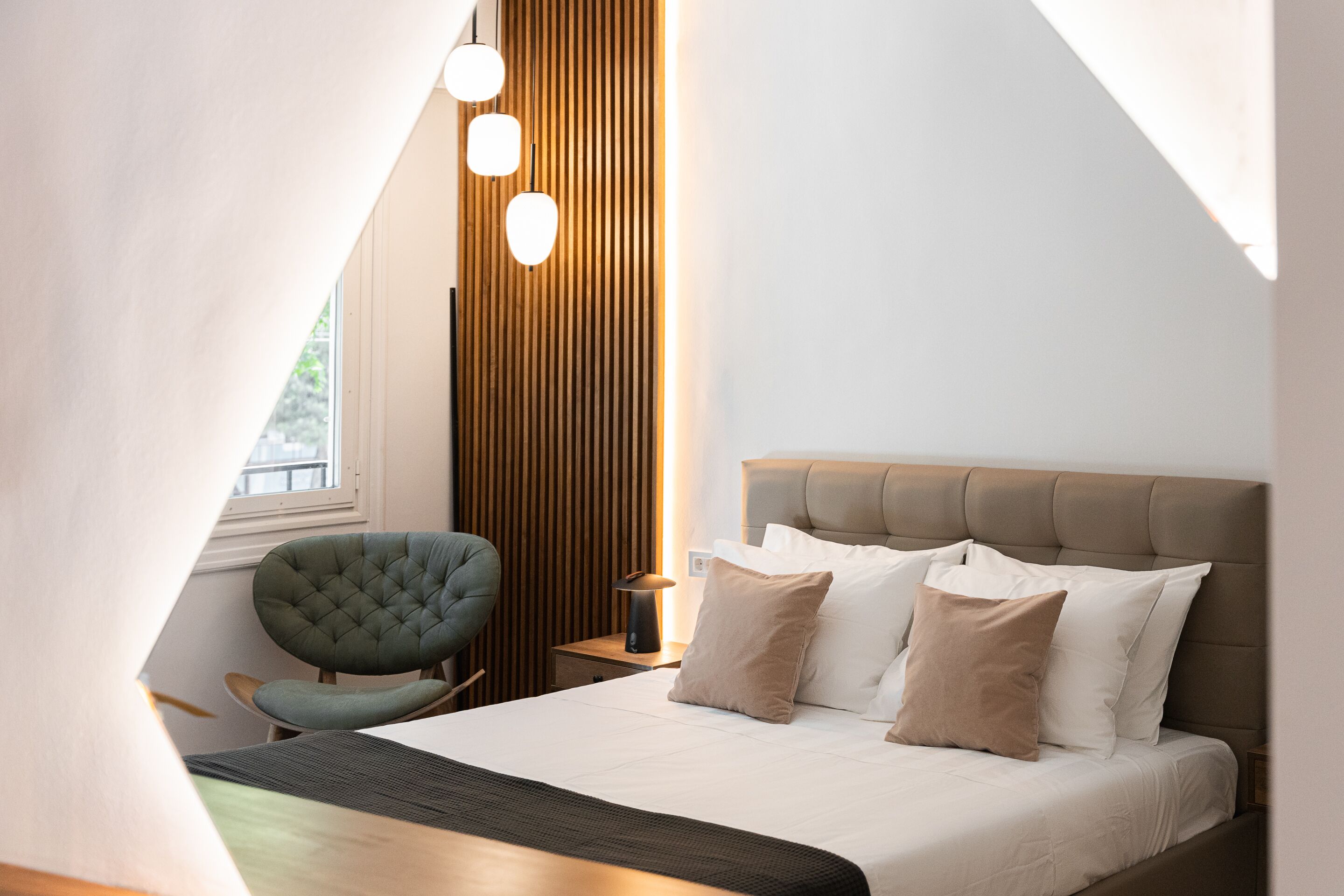 A modern bedroom with a large bed, a green upholstered chair, a wooden accent wall, and a bedside table with a lamp.