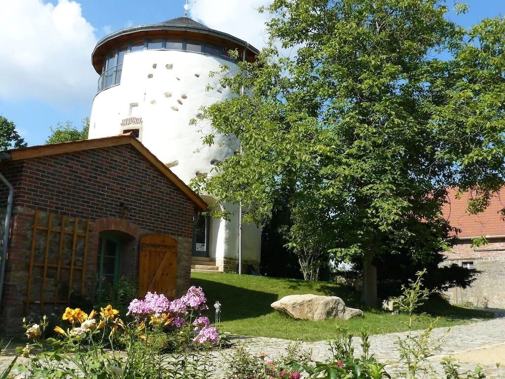 Un edificio con una torre cilíndrica, estructura de ladrillo y un jardín con flores y un árbol.
