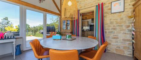 Un coin repas avec une table ronde et des chaises orange, un mur en pierre et une vue sur l’extérieur.