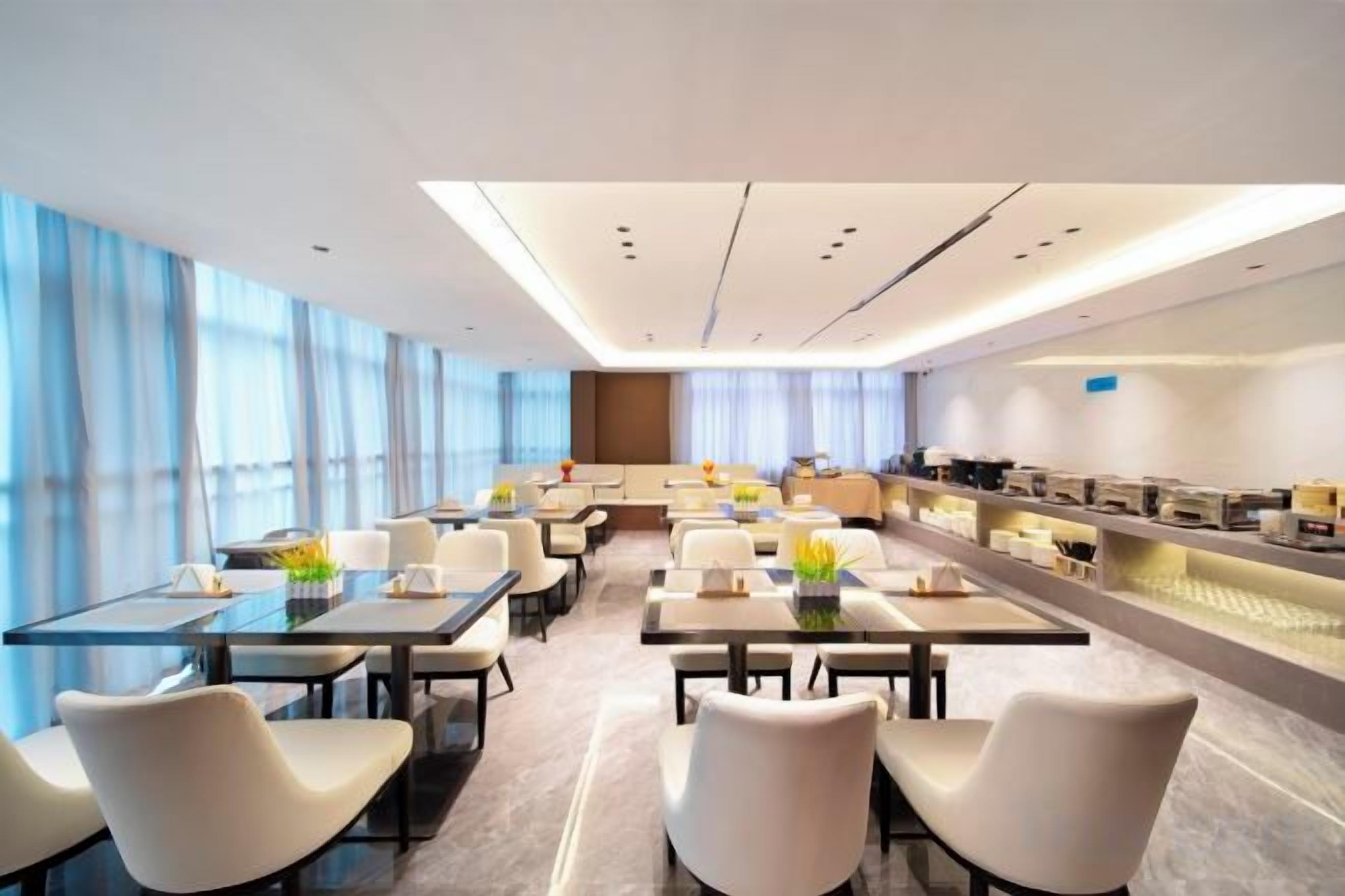 A modern hotel dining area with tables and chairs, a kitchen area, and large windows.