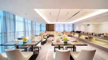 A modern hotel dining area with tables and chairs, a kitchen area, and large windows.