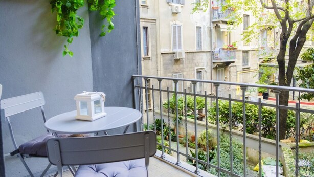 Un balcon avec une table et une chaise, une plante suspendue et une vue sur une cour bordée d’immeubles.