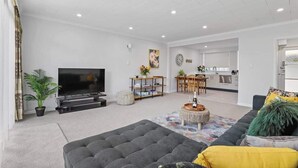 A modern living room with a large grey sofa, a flat-screen TV, a dining area with a wooden table and chairs, and a kitchen in the background.