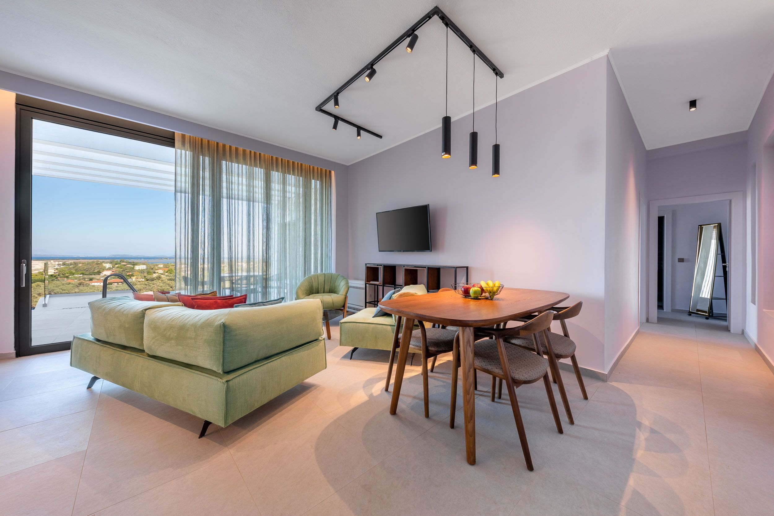 Ein modernes Wohnzimmer mit großem Fenster, einem grünen Sofa, einem Esstisch mit Stühlen und einem an der Wand befestigten Flachbildfernseher.