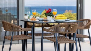 Une table à manger avec des chaises en osier, un plateau en verre et une vue sur la mer grâce à de grandes fenêtres.