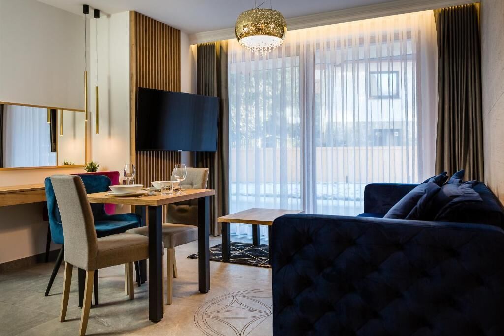 Et moderne hotelværelse med spiseplads, et stort fladskærms-tv, en behagelig sofa og et stort vindue med lette gardiner.