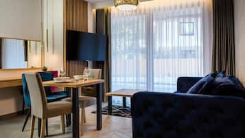 Et moderne hotelværelse med spiseplads, et stort fladskærms-tv, en behagelig sofa og et stort vindue med lette gardiner.