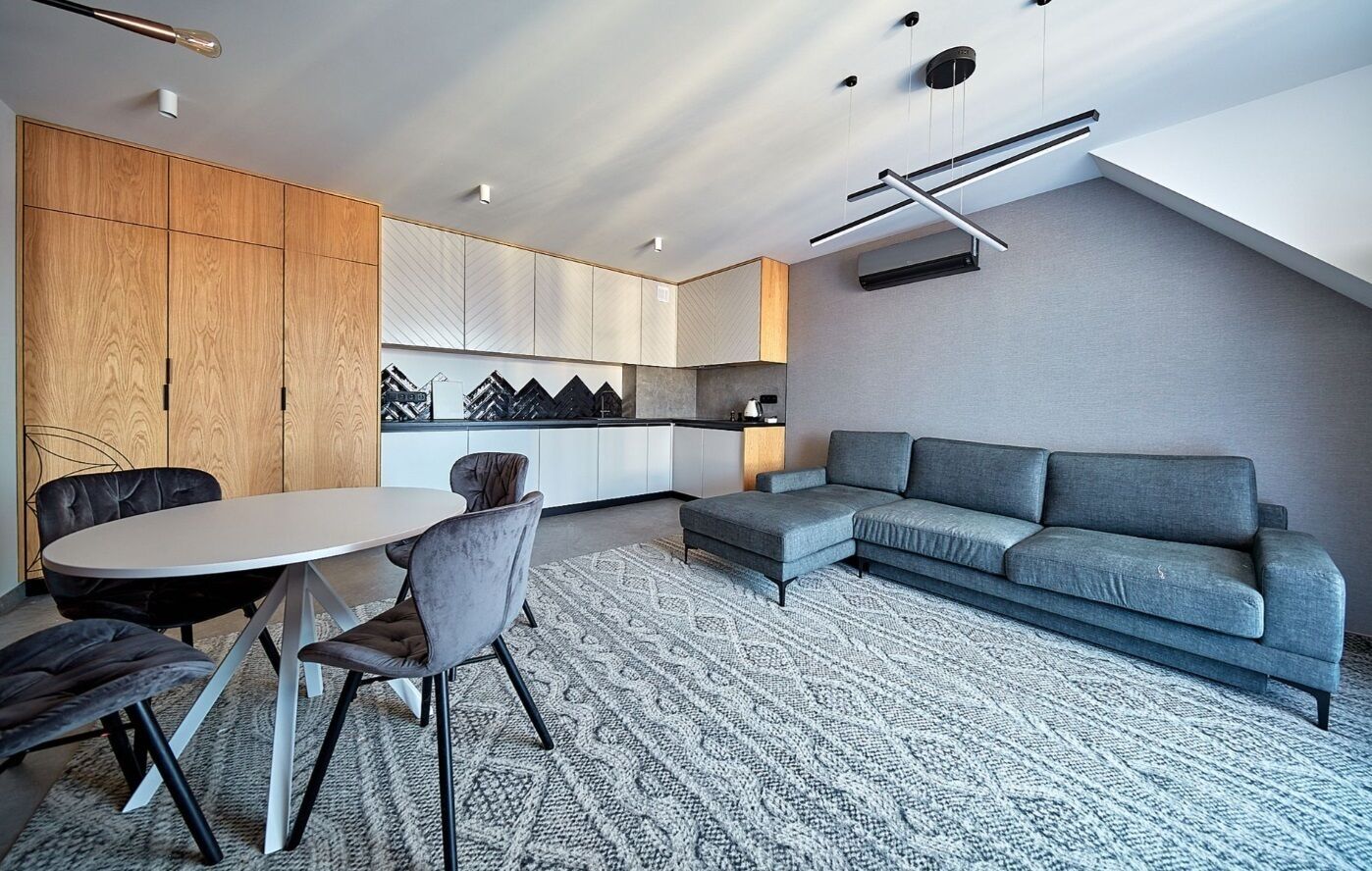Ein modernes Wohnzimmer mit grauem Sofa, einem runden Esstisch mit Stühlen und einer Küchenzeile mit Schränken und einer Arbeitsfläche.