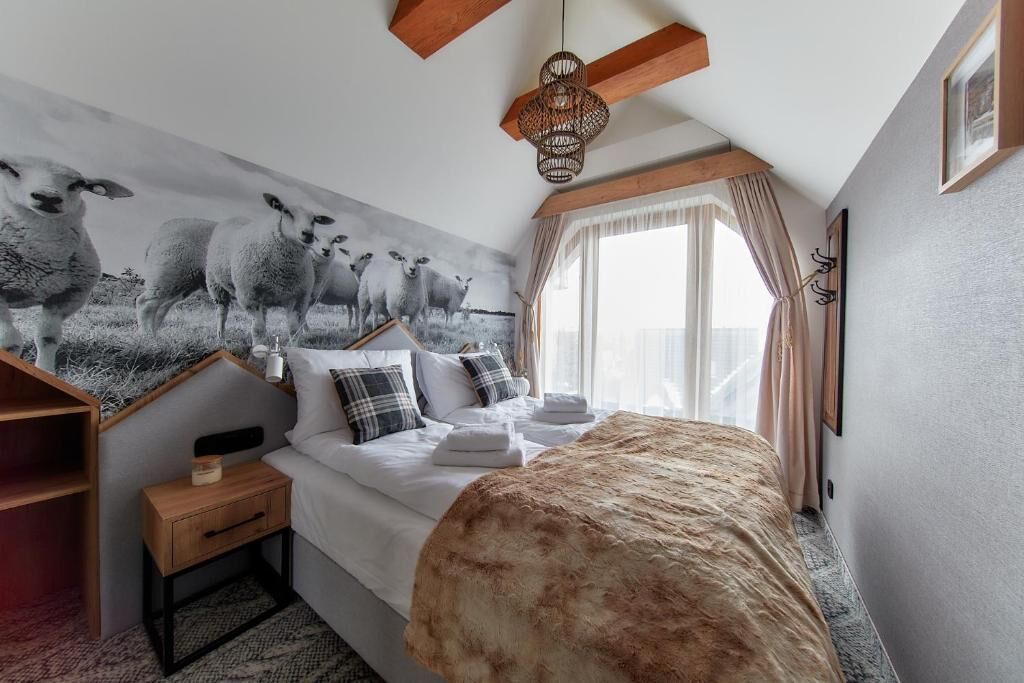 Une chambre à coucher avec un grand lit, une table de chevet, une étagère en bois, un tableau représentant des moutons accroché au mur, un luminaire suspendu et une fenêtre avec des rideaux.